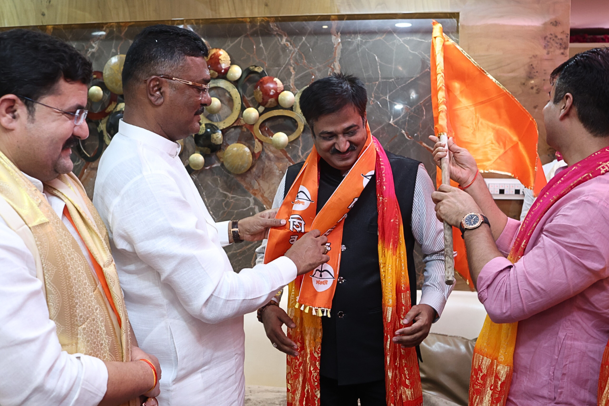 Shiv Sena leader Partap Sarnaik is welcoming Sunil Keshary to the Shiv Sena party by draping an orange party scarf around his neck, with others present and an orange party flag visible.