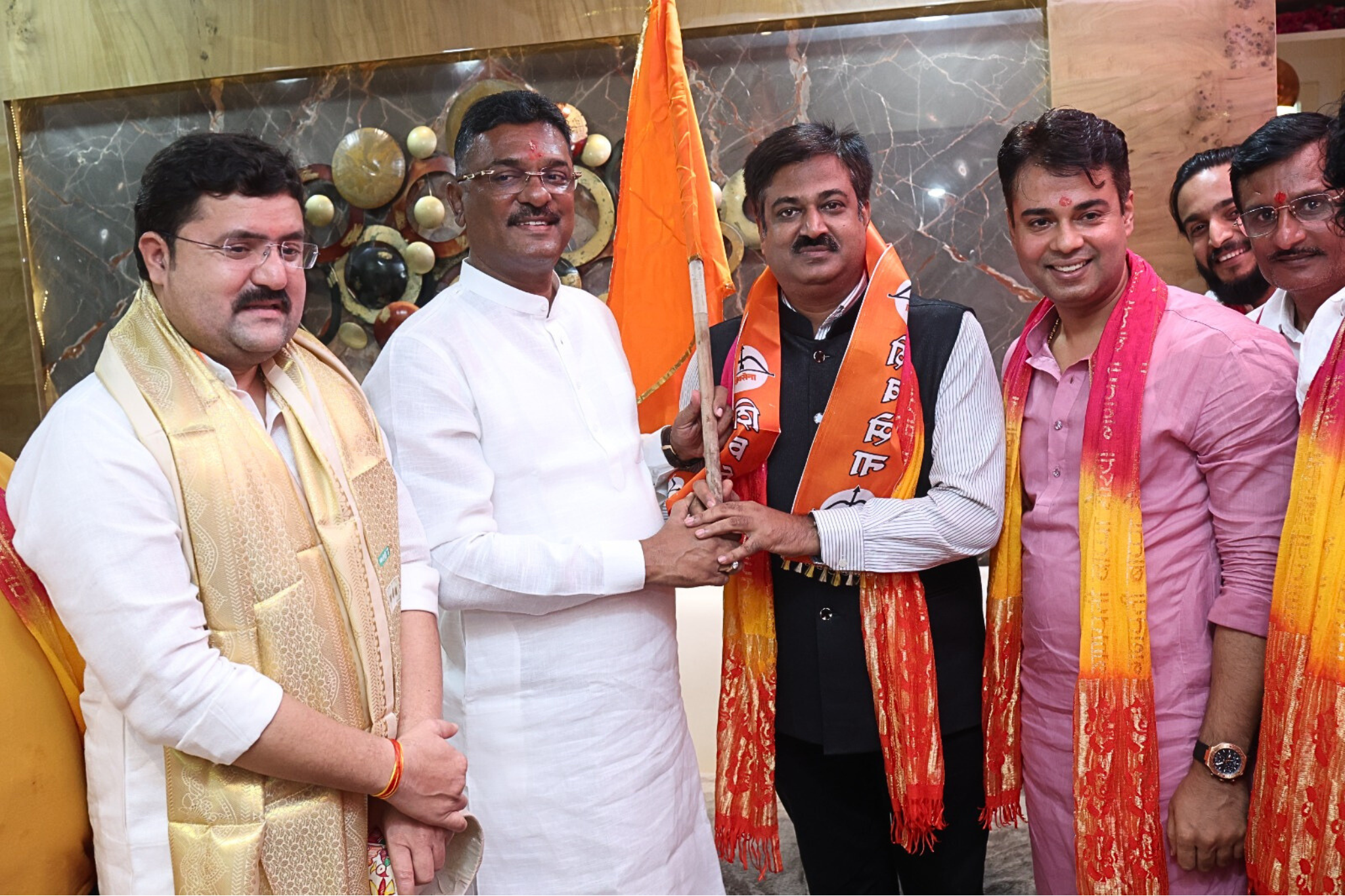 Shiv Sena members, including Sunil Keshary and Partap Sarnaik, are gathered together indoors and jointly raising a small Shiv Sena flag, with both Keshary and Sarnaik holding the flagpole at the center of the group.