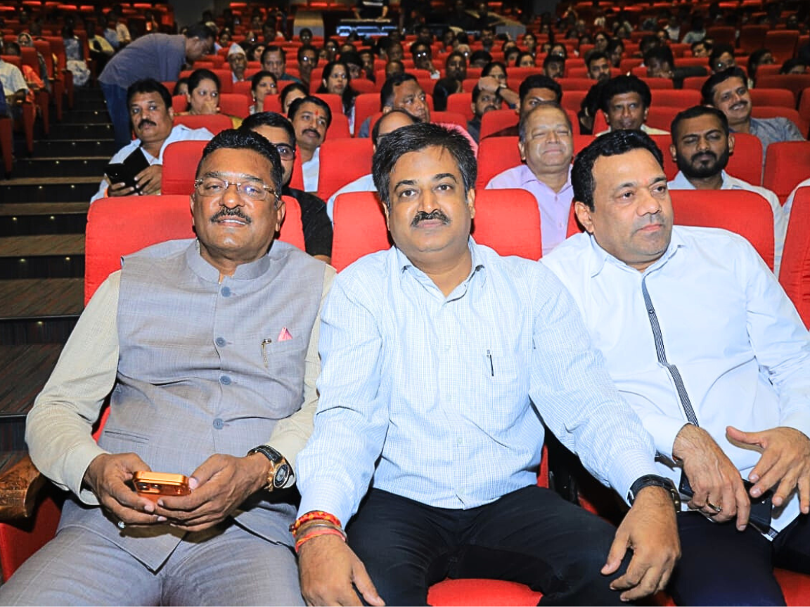 Shiv Sena leaders Partap Sarnaik and Sunil Keshary, along with another dignitary, are seated in the front row of a large auditorium with tiered red seating, attending a public meeting, cultural program, or political rally. The image captures the leaders observing the event with the crowd, while one attendee attempts to capture a photo on their phone.