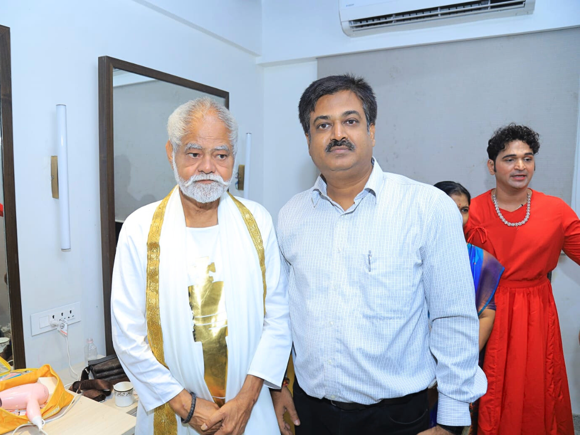 Shiv Sena leader Partap Sarnaik is meeting with a distinguished, white-bearded artist dressed in white robes and a gold sash, likely in a backstage or dressing room area of an auditorium. The photo suggests Sarnaik is greeting the artist after a cultural performance.