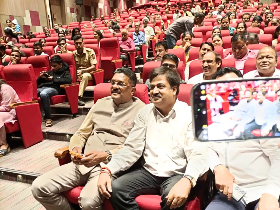 Shiv Sena leaders Partap Sarnaik and Sunil Keshary, along with another dignitary, are seated in the front row of a large auditorium with tiered red seating, attending a public meeting, cultural program, or political rally. The image captures the leaders observing the event with the crowd, while one attendee attempts to capture a photo on their phone.
