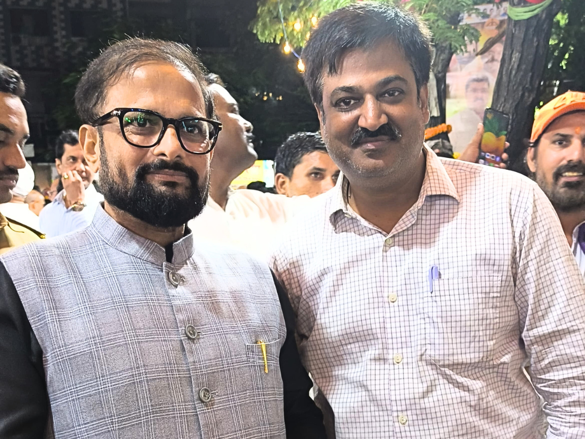 Shiv Sena leader Partap Sarnaik is smiling while posing closely with another party or community official during a nighttime public event or rally. The setting is outdoors with crowds and promotional banners visible in the blurred background.