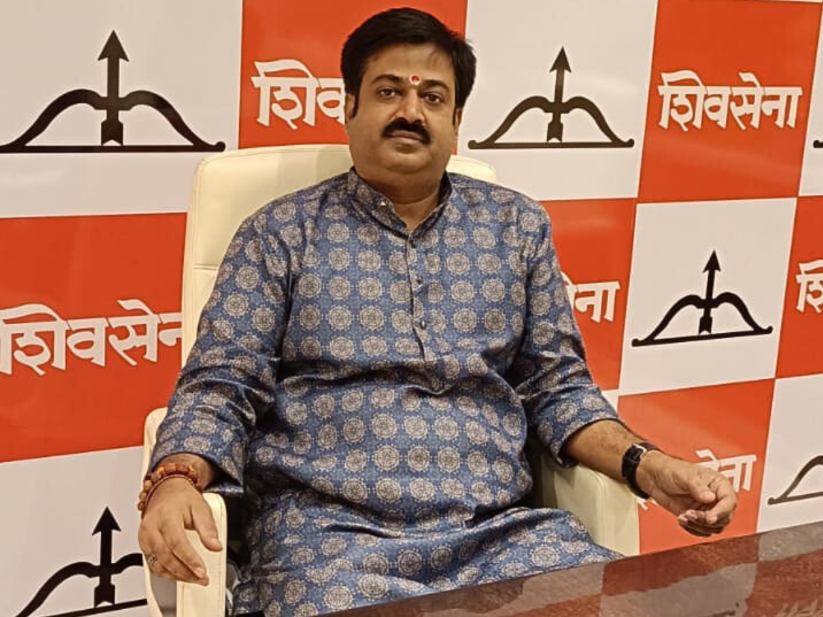 Sunil Keshary is seated in a white chair, wearing a blue patterned kurta and crossing his legs, in front of a Shiv Sena logo backdrop featuring the bow-and-arrow symbol and party name.