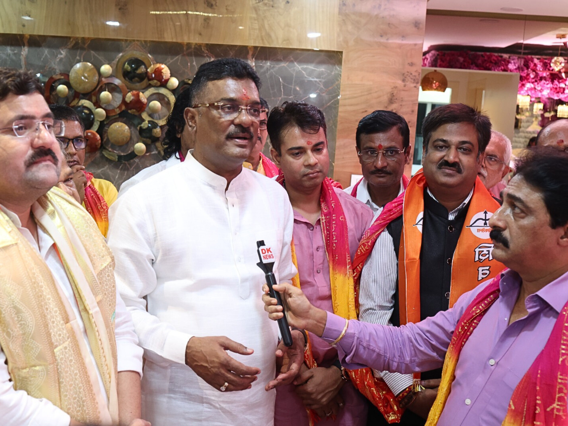 Sunil Keshary stands with multiple people during an indoor event, as Partap Sarnaik, dressed in white, is being interviewed by someone holding a microphone; others wear festive orange scarves and are gathered for a group interaction.