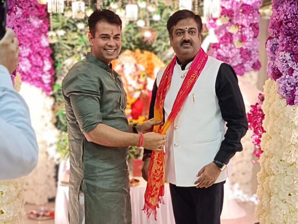 Sankar Jha is putting a festive scarf around Sunil Keshary, both standing together in front of a decorative Ganpati setup with vibrant flowers and an idol in the background.